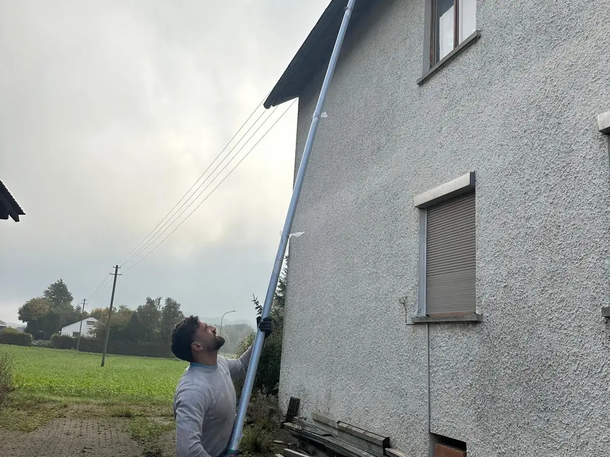 Ein Mitarbeiter führt eine ausgefahrene Teleskopstange vom Boden aus an die Dachrinne eines Wohnhauses.