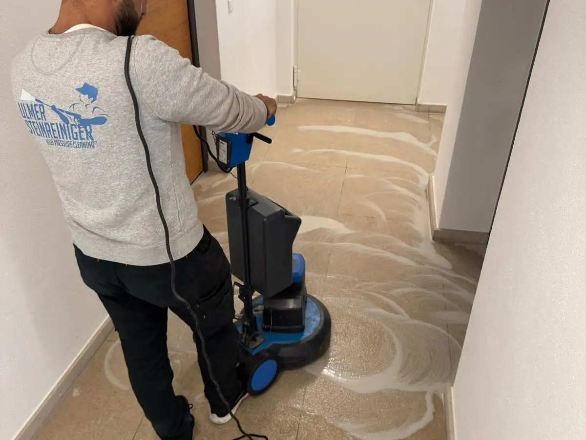 Ein Mitarbeiter führt eine Einscheibenmaschine über den Fliesenboden eines Treppenhauses, auf dem Belag steht Reinigungsschaum.