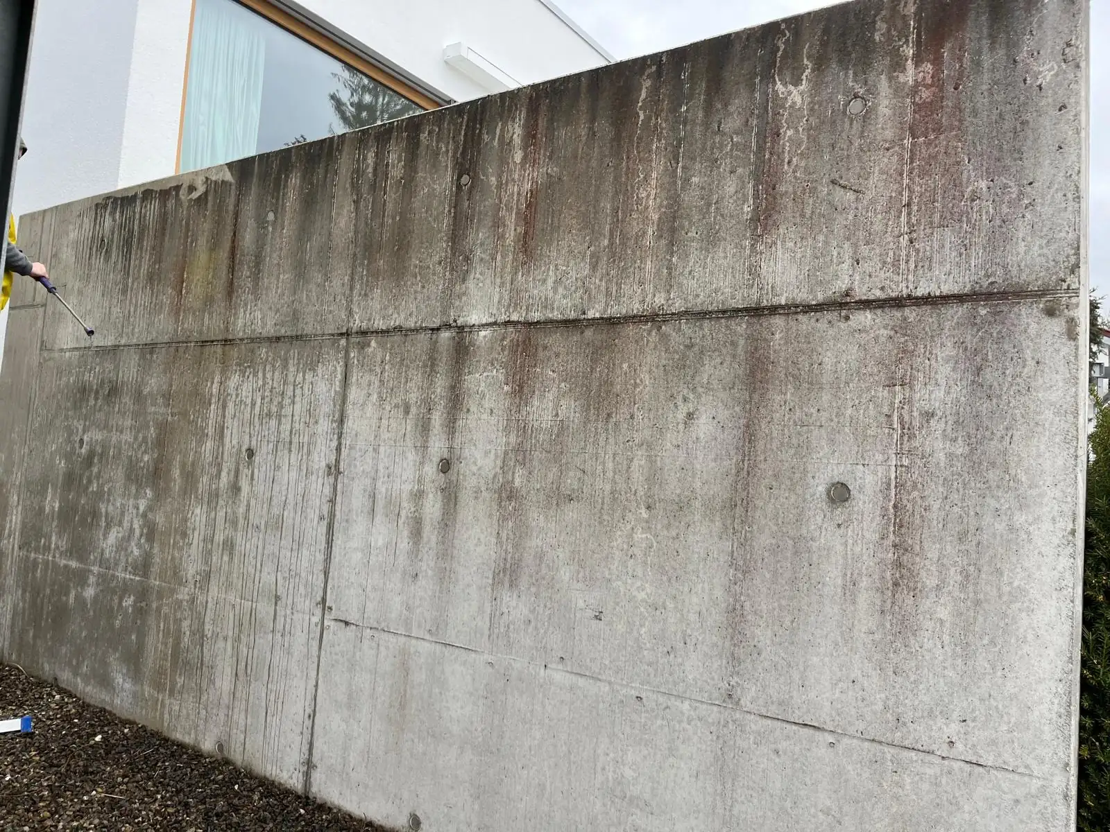 Sichtbetonmauer an einem Wohnhaus, links im ungereinigten Zustand mit braunen Ablaufspuren, rechts nach der Reinigung gleichmäßig grau., vor der Reinigung